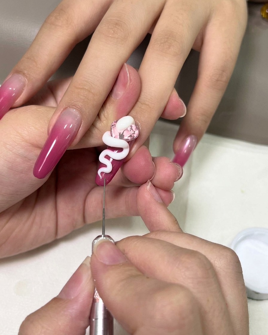 Nahaufnahme einer Hand, die mit einem feinen Metallwerkzeug eine weiße 3D-Gelschlange über einem rosa Edelstein auf einem magentafarbenen Ombré-Nagel in Form bringt.