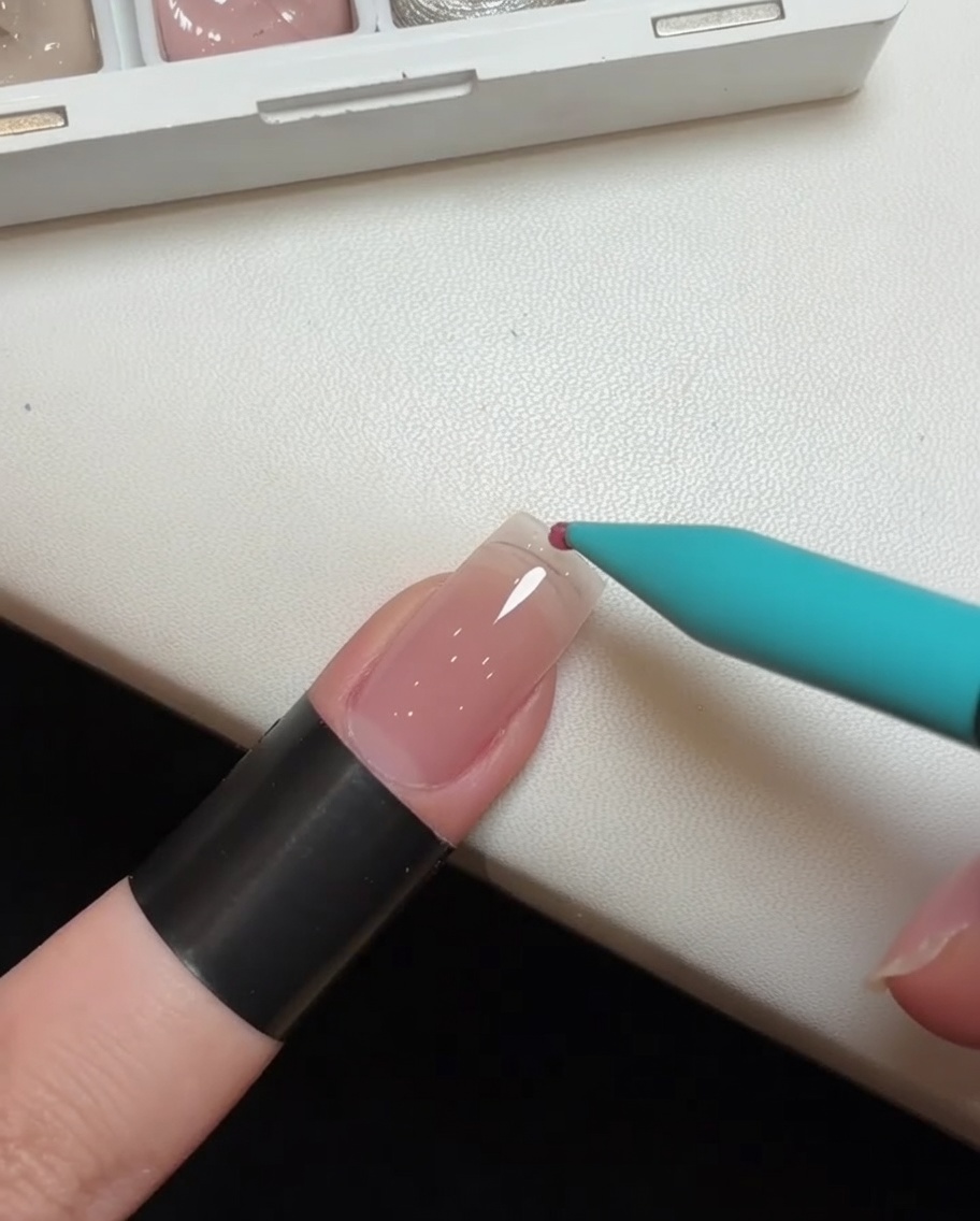 Nahaufnahme eines Detailpinsels, der einen Tropfen rosa Gellack auf die Spitze einer transparenten Nagelverlängerung aufträgt.