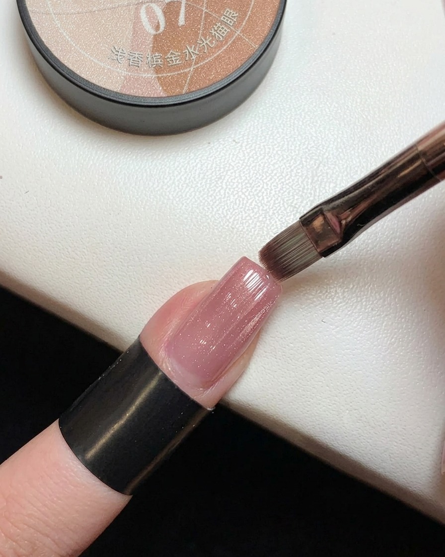 Ein Pinsel trägt ein halbtransparentes, rosa Magnetgel mit Goldschimmer über einem zuvor ausgehärteten rosa Ombré-Nagel auf.