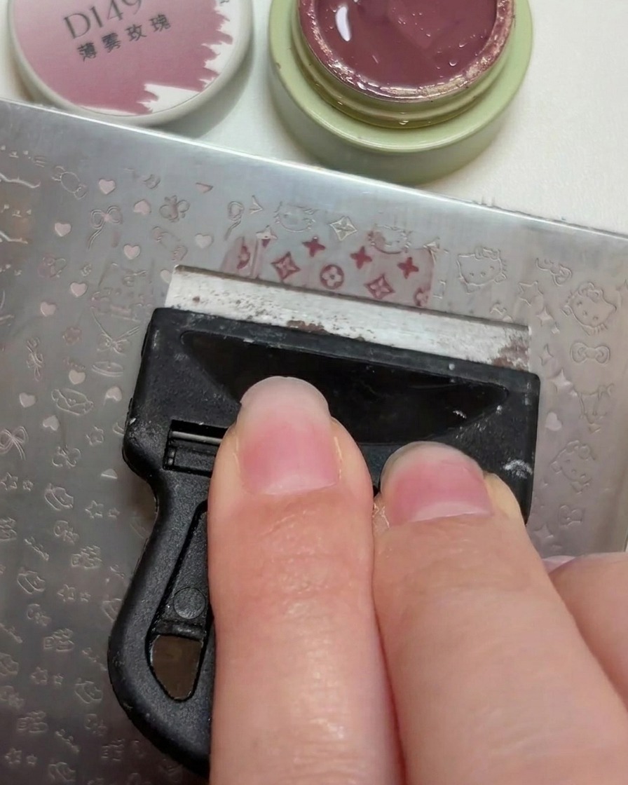 Ein Plastik-Scraper zieht einen kleinen Klecks altrosafarbenen Gellack über die eingravierten Muster auf einer Nagel-Stamping-Schablone ab.