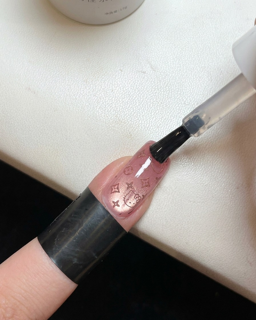Ein Pinsel trägt einen klaren, glänzenden Top Coat über einem Nagel mit rosa Hello Kitty Muster auf.