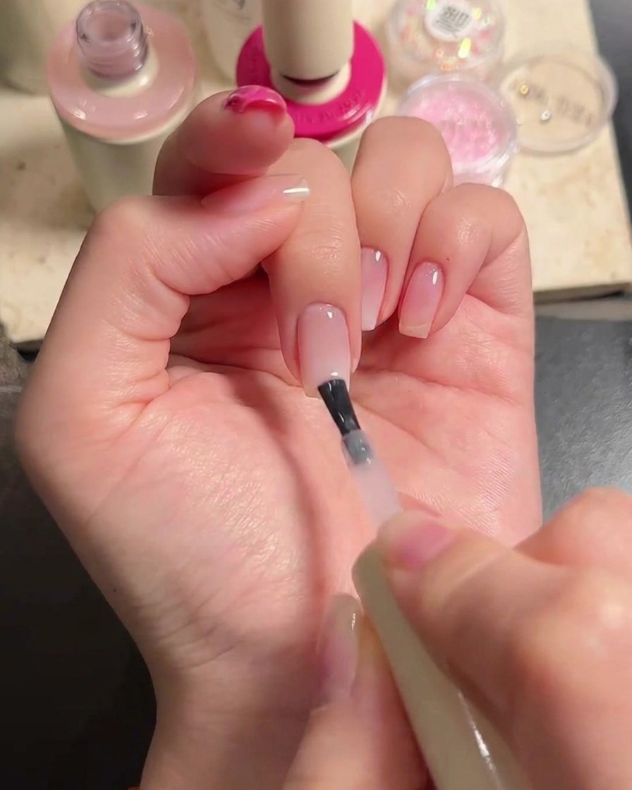 Ein Pinsel trägt eine Schicht transparentes Nude-Gel über Mittel- und Ringfinger auf, um den Verlauf zu verblenden.
