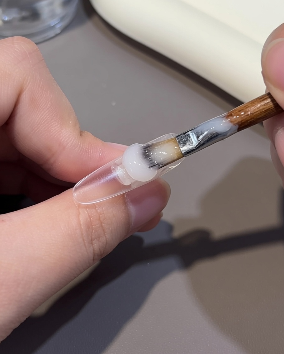 Nahaufnahme eines Pinsels, der ein nudefarbenes Gel auf den hinteren Teil eines klaren Nagel-Tips aufträgt.