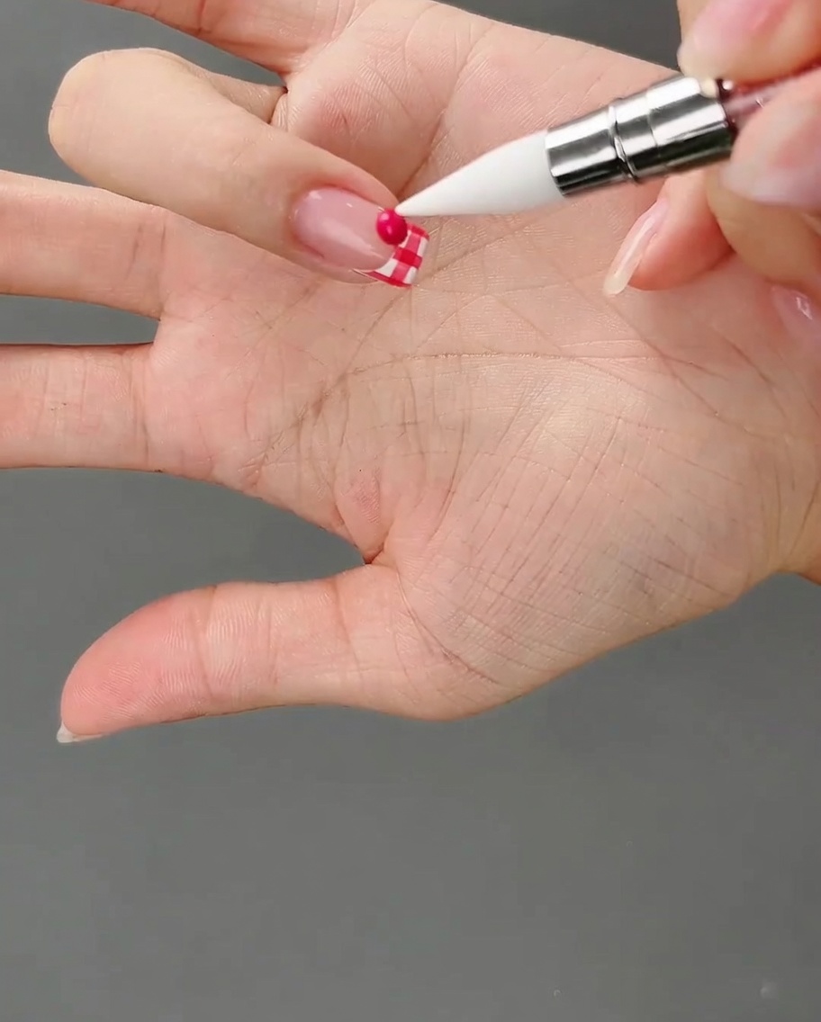 Ein Silikonwerkzeug zeigt auf ein kleines, rundes Kügelchen aus dunklem Pink-Gel auf dem karierten Nagel.
