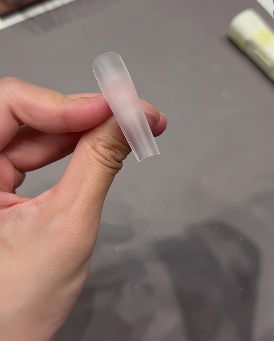 Eine Hand platziert einen transparenten Klebepad auf die Oberfläche eines klaren Nagel-Tips.