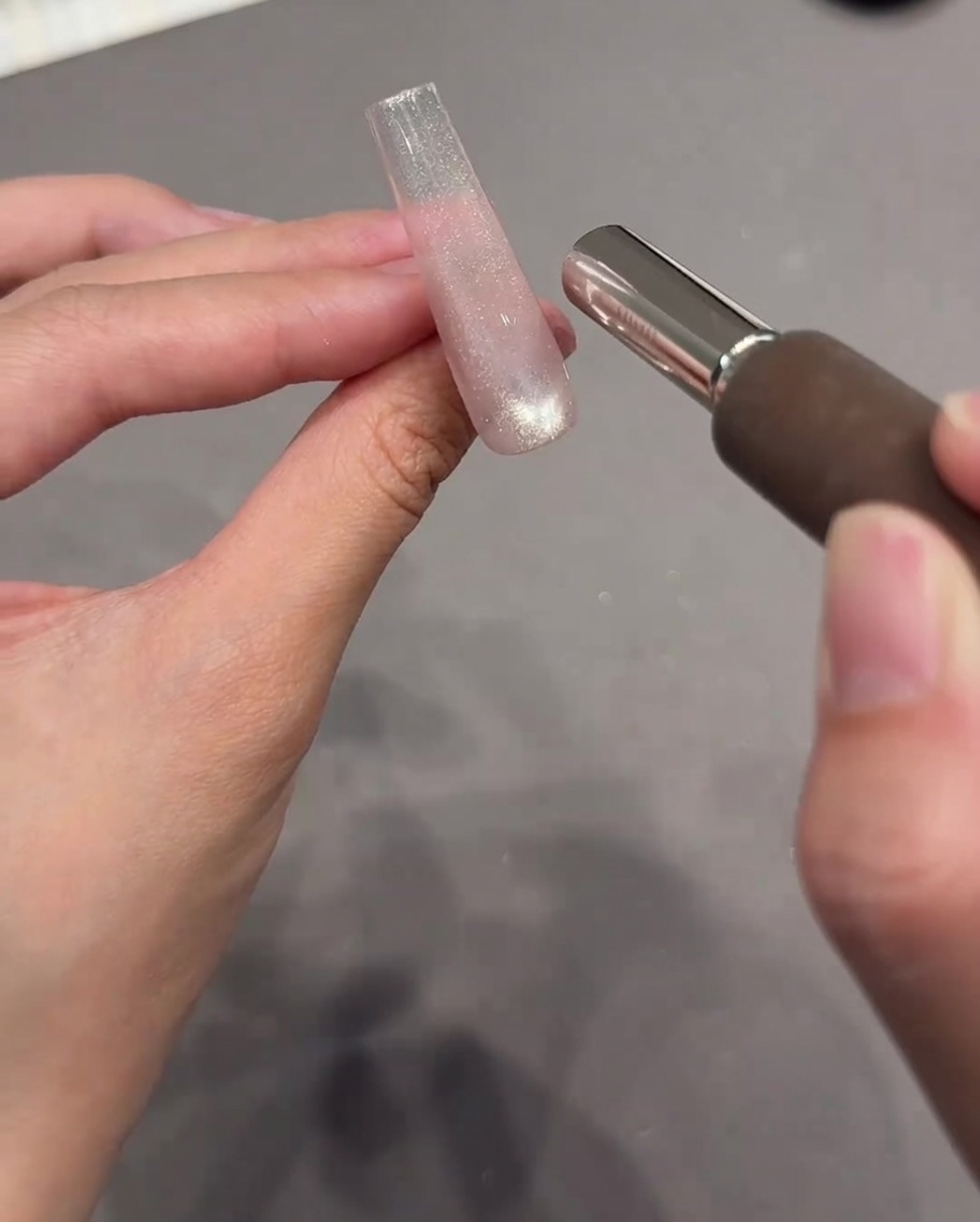 Eine Hand hält ein zylindrisches Magnetwerkzeug nahe an eine klare Nagelverlängerung mit feuchtem, glitzerndem Cat-Eye-Gel.