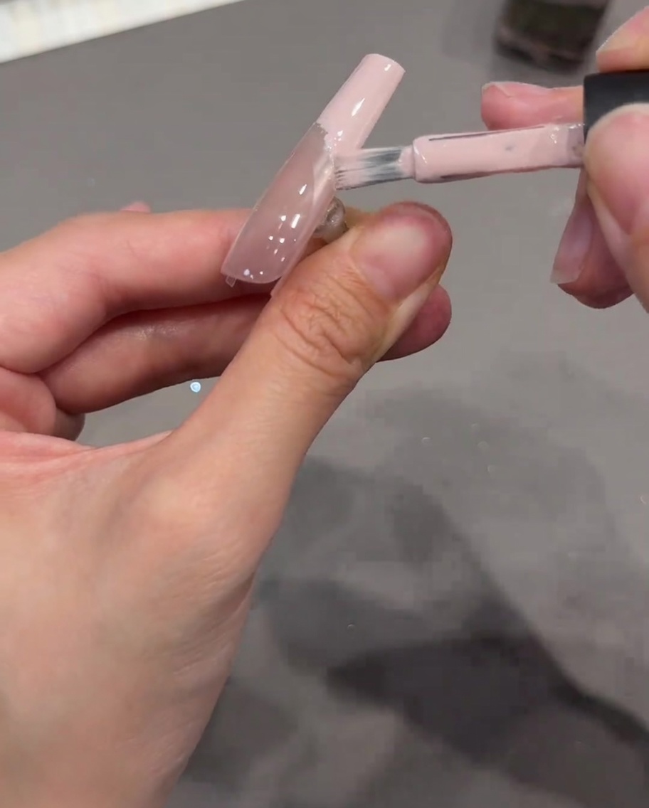 Ein Nagelpinsel trägt deckendes nudefarbenes rosa Gel auf die Spitze eines Nagels auf, wobei ein klarer Aufkleber als Grenze dient.