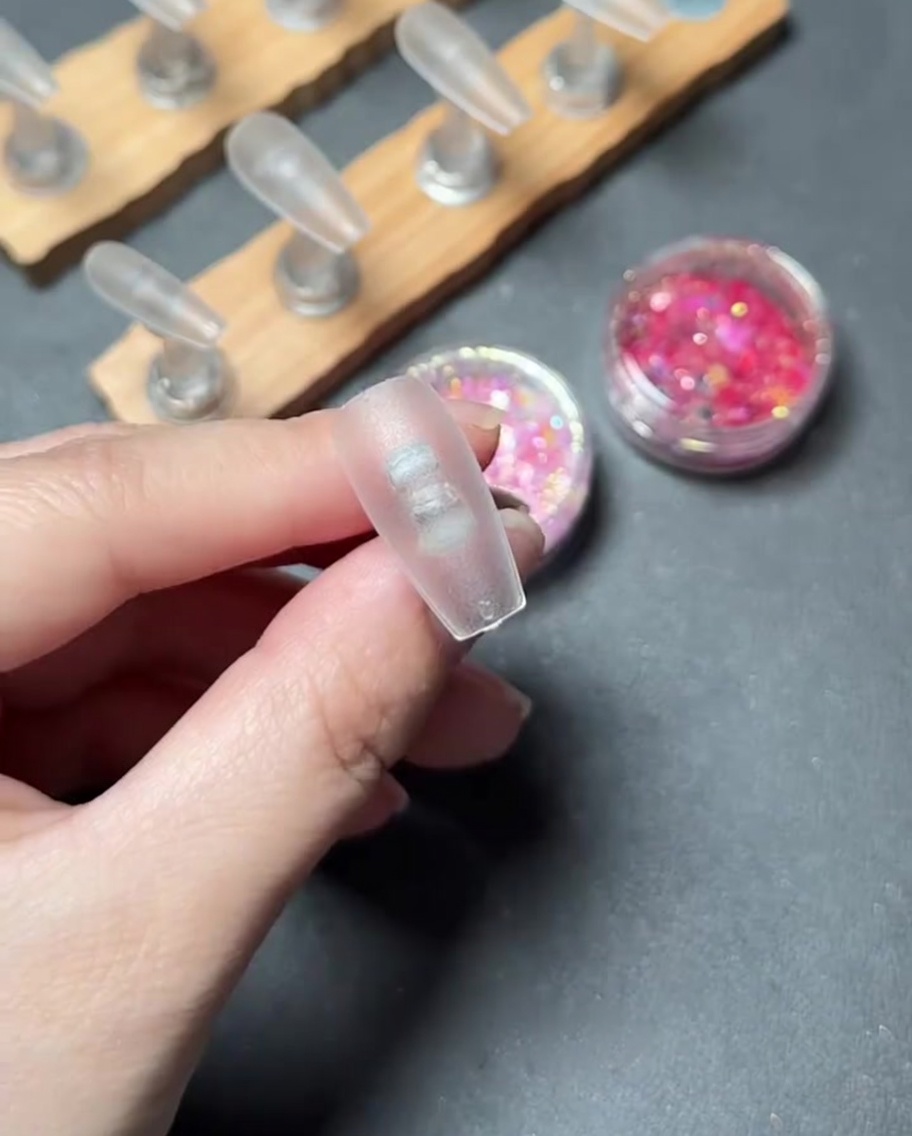 Nahaufnahme eines Pinsels, der klares Basis-Gel auf die Oberfläche eines durchsichtigen Kunststoff-Nageltips aufträgt.