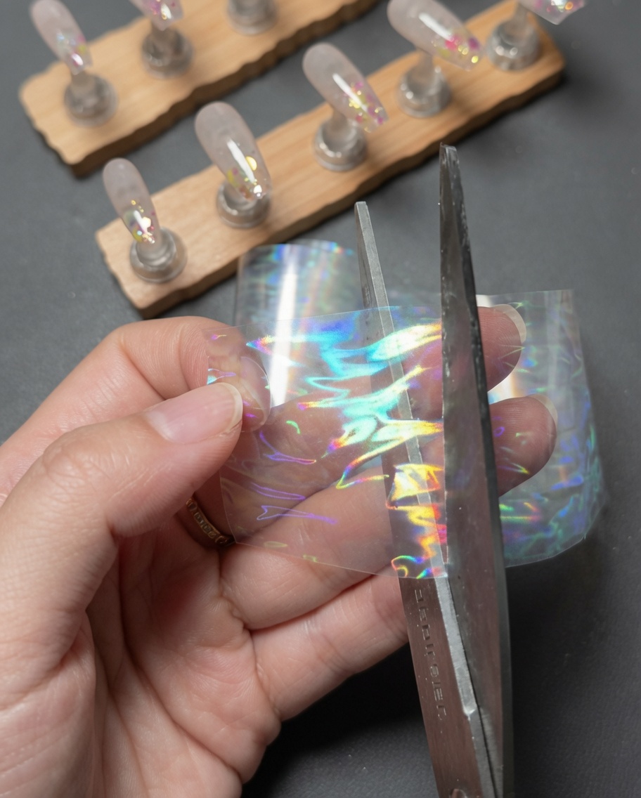 Hände, die mit einer Schere ein kleines rechteckiges Stück von einem Streifen holografischer Laser-Aurora-Transferfolie abschneiden.