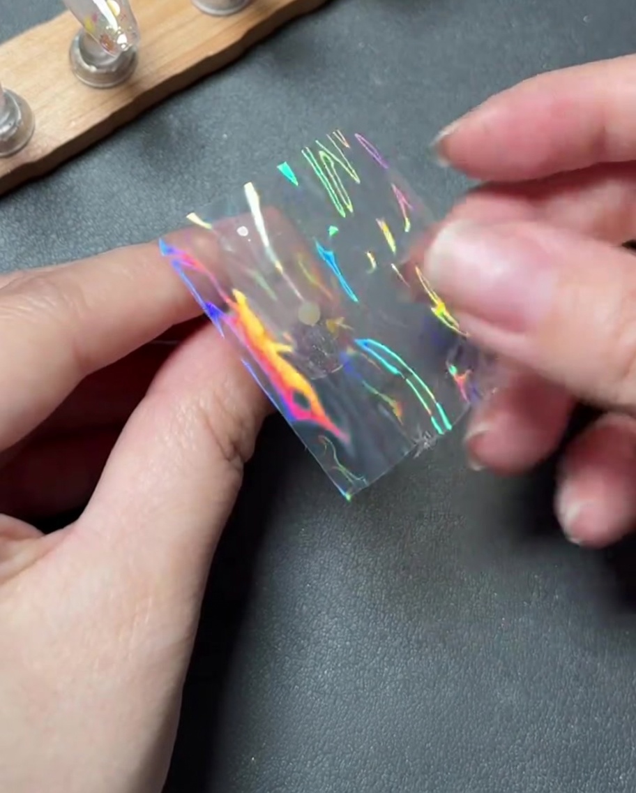 Finger, die vorsichtig ein kleines Stück holografische Transferfolie in die Mitte eines Nagels legen.