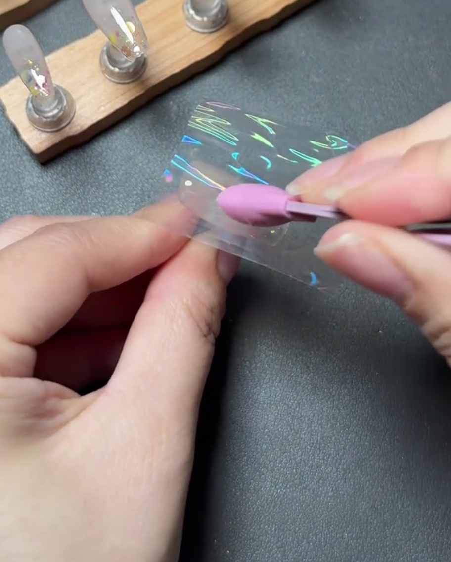 Ein rosa Silikonwerkzeug, das ein Stück holografische Transferfolie fest auf einen Nagel drückt und glättet.