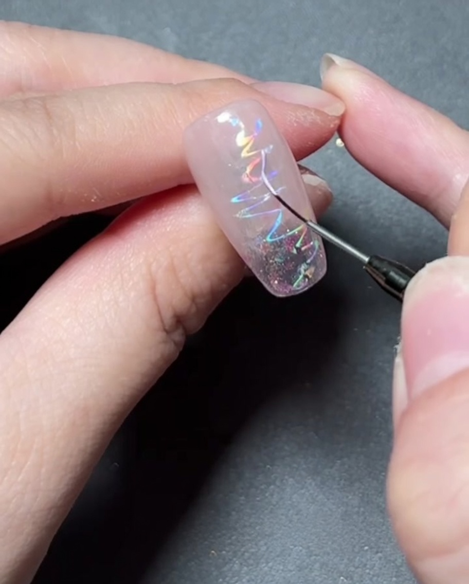 Ein feiner Liner-Pinsel, der mit buntem Gel ein zartes Sternmotiv auf einen holografisch folierten Nagel malt.