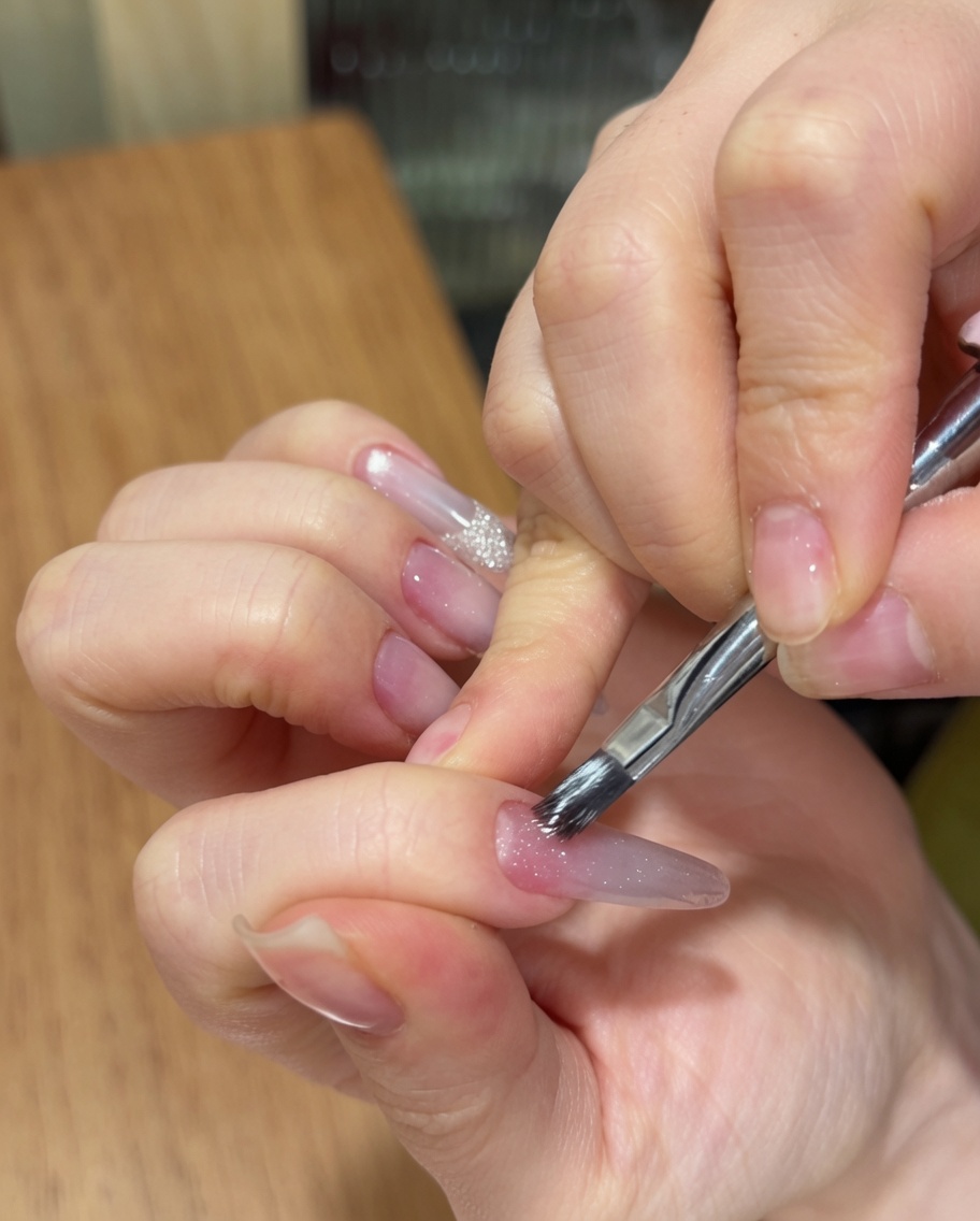 Eine Hand trägt mit einem Nagelpinsel ein funkelndes Cat-Eye-Gel über einen langen mandelförmigen Nagel mit rosafarbenem Ombré-Verlauf auf.