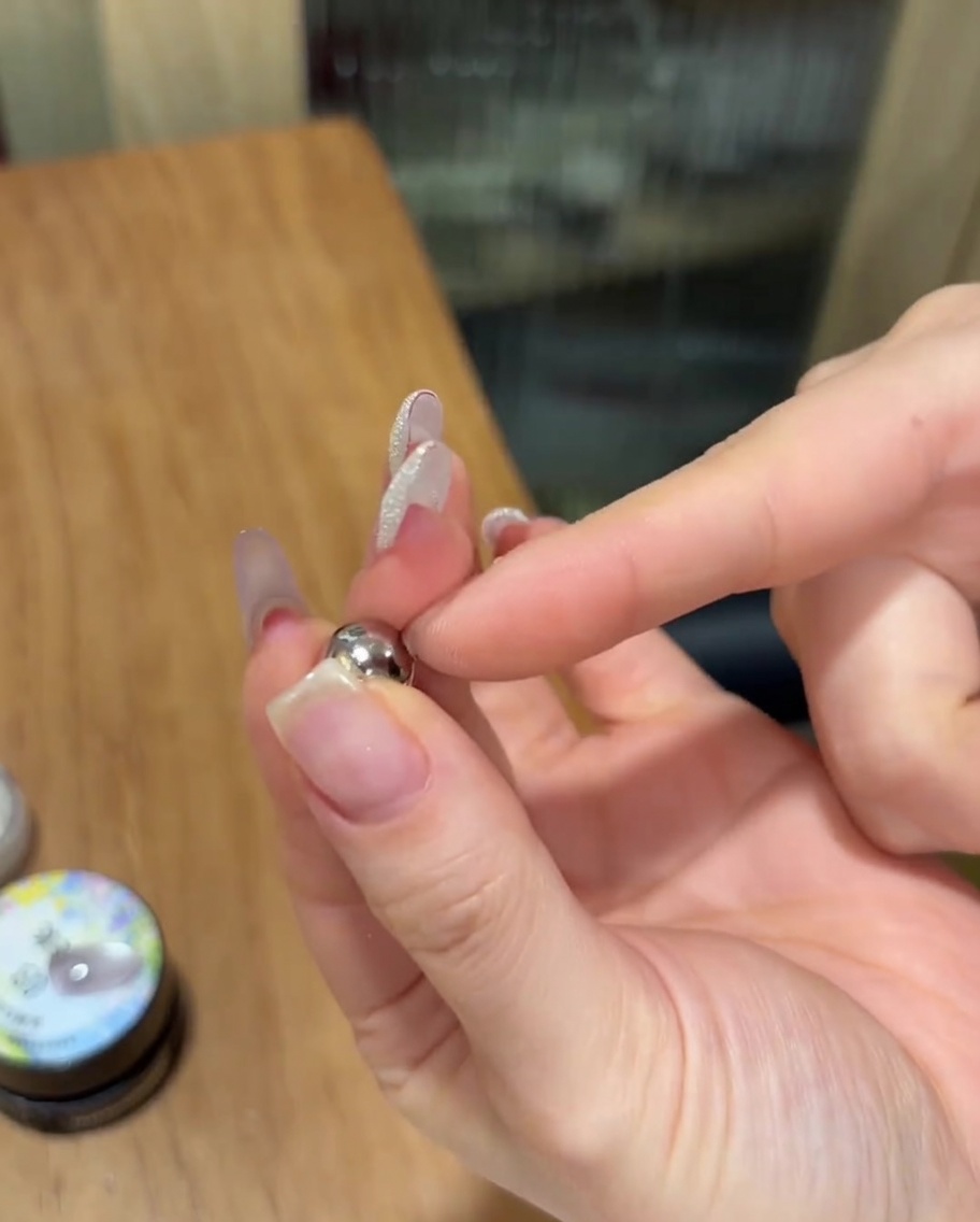 Eine Hand hält einen kleinen kugelförmigen, silbernen Magneten dicht über einen Fingernagel, der mit noch feuchtem Cat-Eye-Gel lackiert ist.