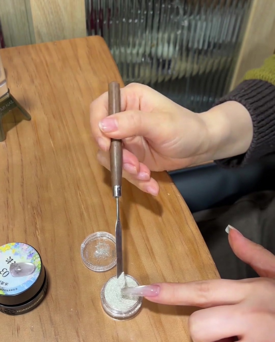 Nahaufnahme eines Nagels, bei dem feuchtes klares Gel mit einem Metallspatel reichlich mit weißem Sugar Powder bestreut wird.