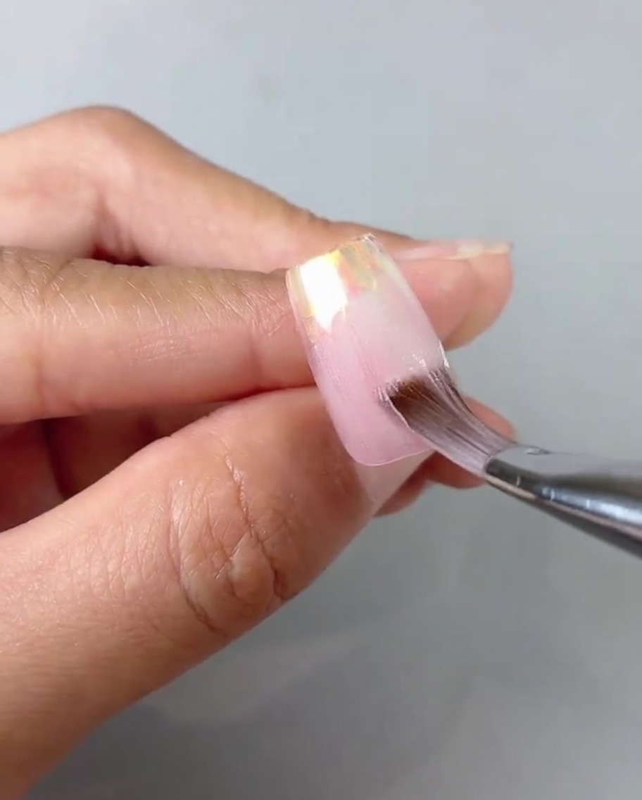 Nahaufnahme eines Pinsels, der transparentes Pink-Gel auf einem Nageltip glättet.