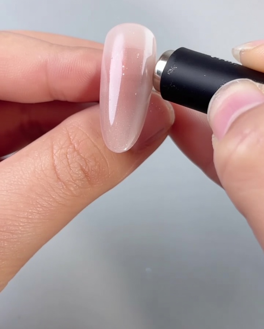Eine Hand hält einen zylindrischen Magneten dicht über einen mandelförmigen Nagel und aktiviert die schimmernden Partikel im nassen Cat-Eye-Gel.
