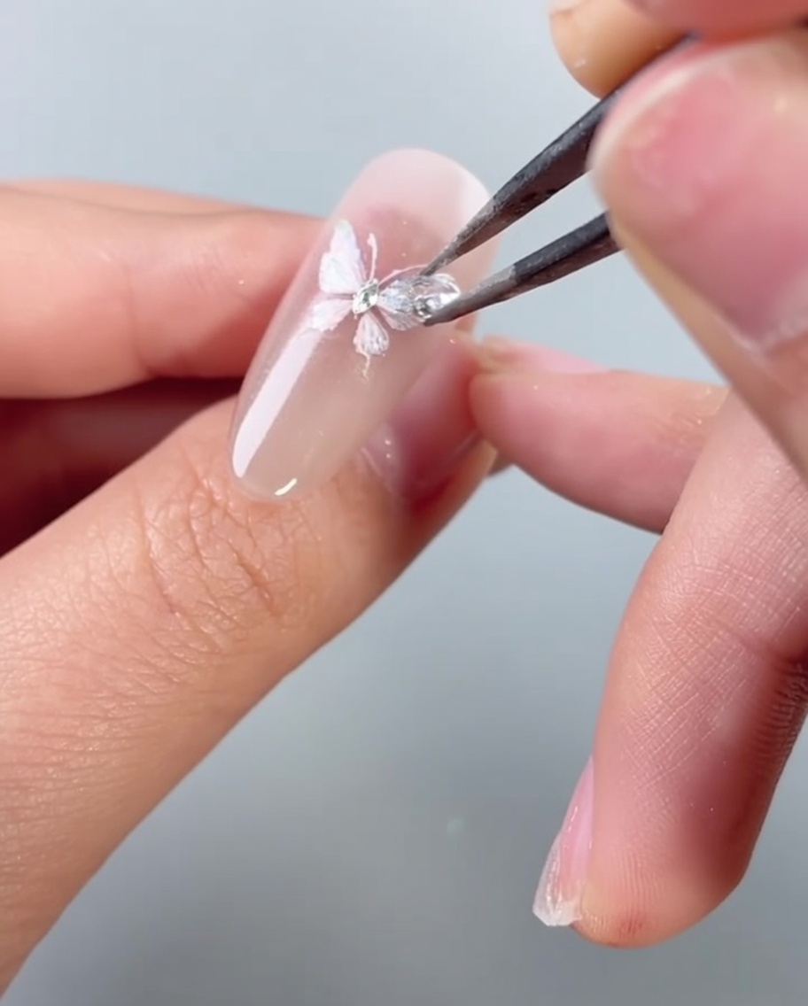 Eine Pinzette platziert einen kleinen, schimmernden Strassstein auf einem verzierten Schmetterlings-Nageldesign.