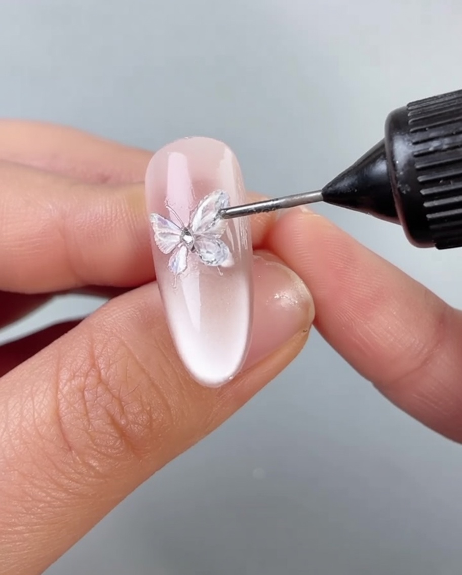 Ein Pinsel trägt eine dünne Schicht klares Gel um die Basis einer Schmetterlingsdekoration auf einem Nagel auf.
