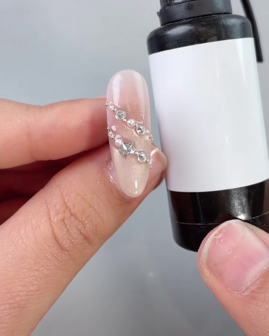 Ein Nagelpinsel trägt einen klaren Top Coat oder eine Versiegelung über einen mit Perlen und Strasssteinen verzierten Nagel auf.