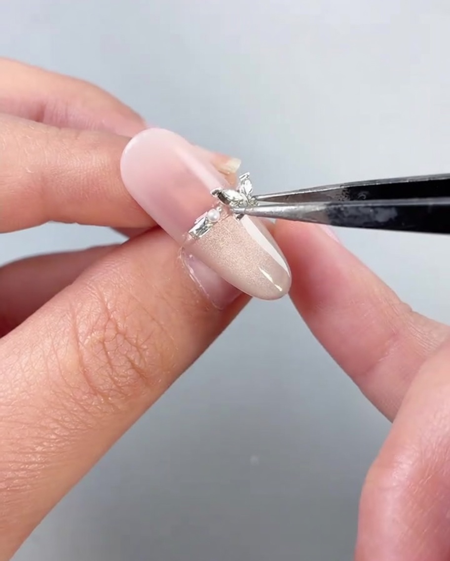 Eine Pinzette platziert einen metallischen Schmetterlings-Charm auf einem verzierten Fingernagel.