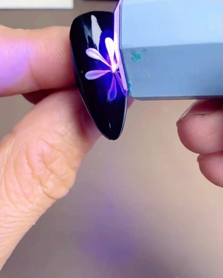 Eine kleine Hand-UV/LED-Lampe schwebt über einer Nagelspitze mit einem weiß-gelben Gänseblümchen-Design.