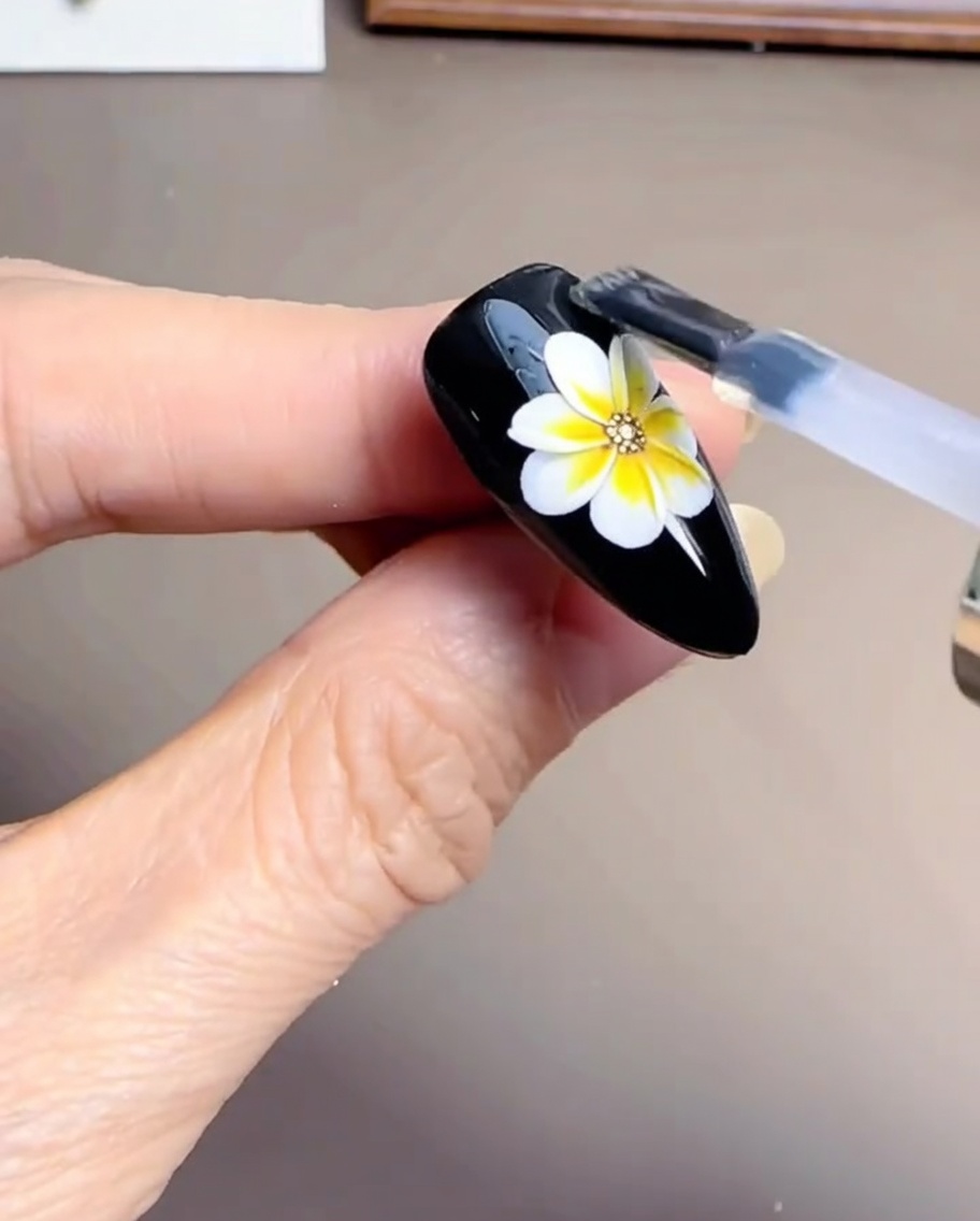 Ein Nagelpinsel trägt eine klare, glänzende Versiegelung über ein teilweise fertiges Gänseblümchen auf einem schwarzen Nagel auf.