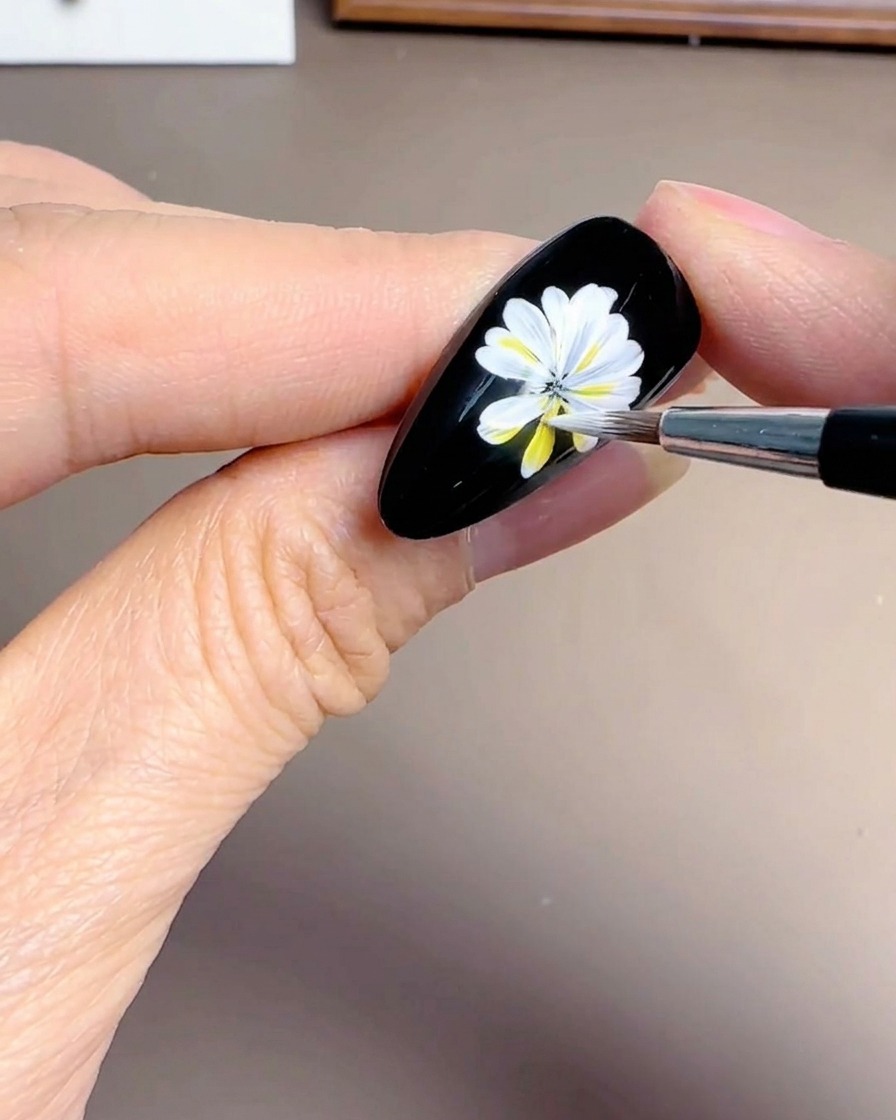 Nahaufnahme einer Hand, die überlappende weiße Blütenblätter zu einem Gänseblümchen-Design auf einem schwarzen Nagel hinzufügt.