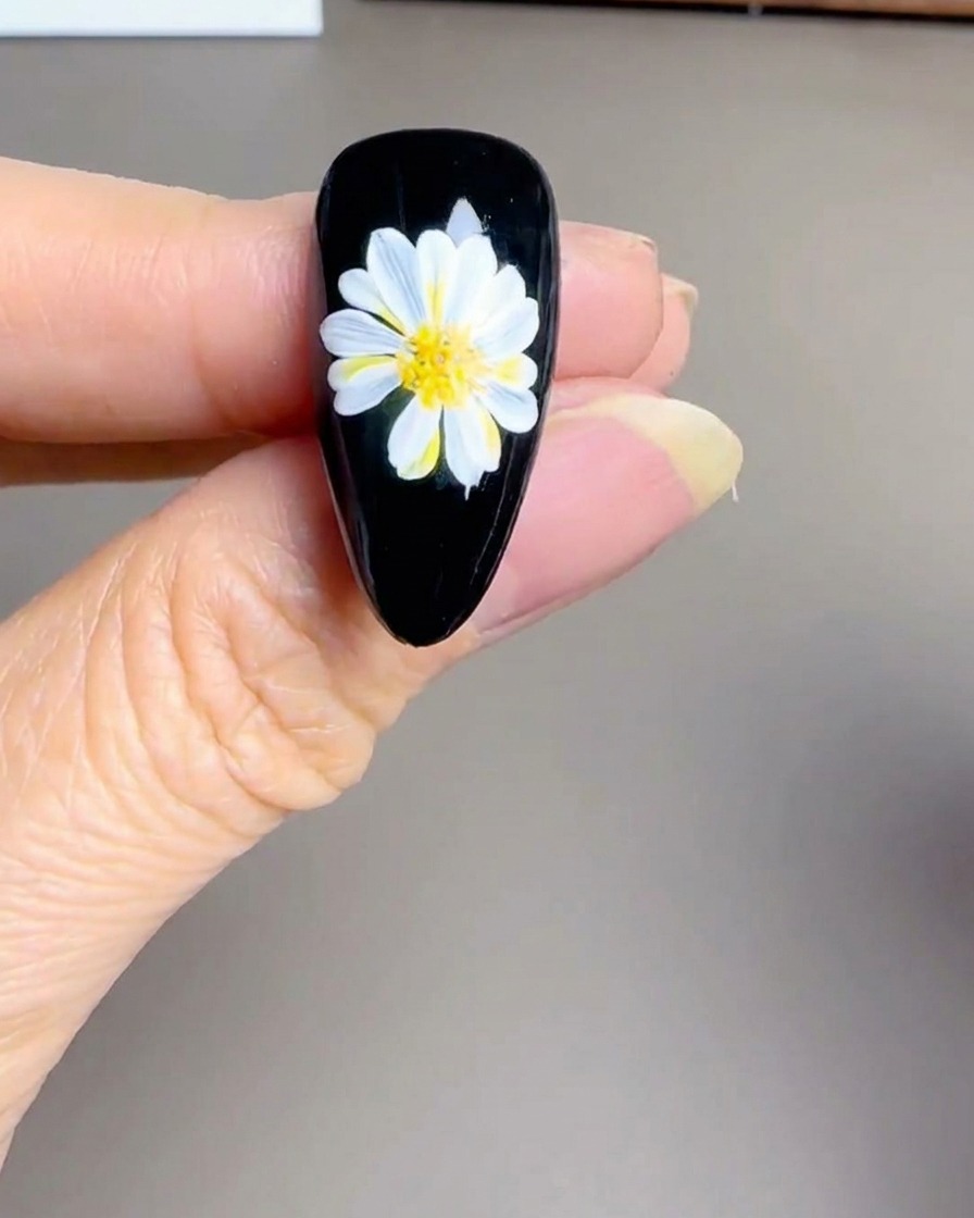 Nahaufnahme einer Hand, die eine kleine UV/LED-Lampe über einen fertigen schwarzen Nagel mit Gänseblümchen-Design hält.