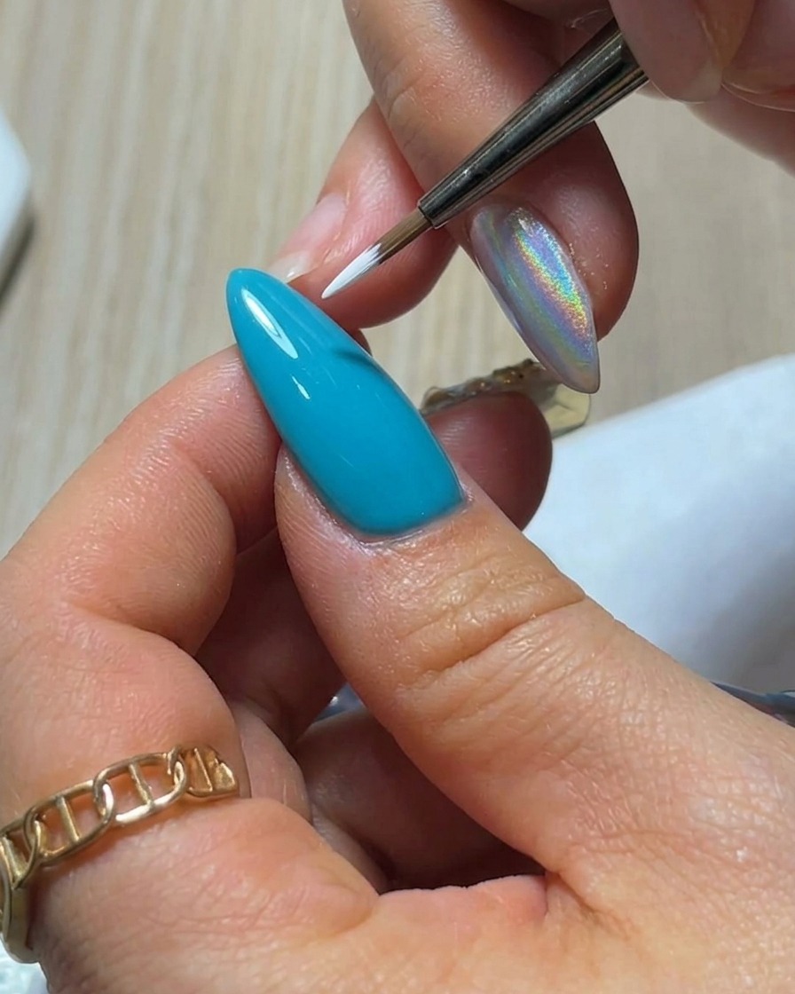 Eine Nahaufnahme eines feinen Liner-Pinsels, der ein einzelnes, tropfenförmiges weißes Gel-Blütenblatt auf eine leuchtend blaue Nageloberfläche aufträgt.
