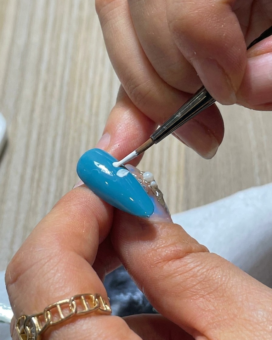 Eine Nahaufnahme zeigt, wie ein drittes weißes Blütenblatt zum blauen Nagel hinzugefügt wird, womit ein Teil des äußeren Rings des Gänseblümchens vervollständigt wird.