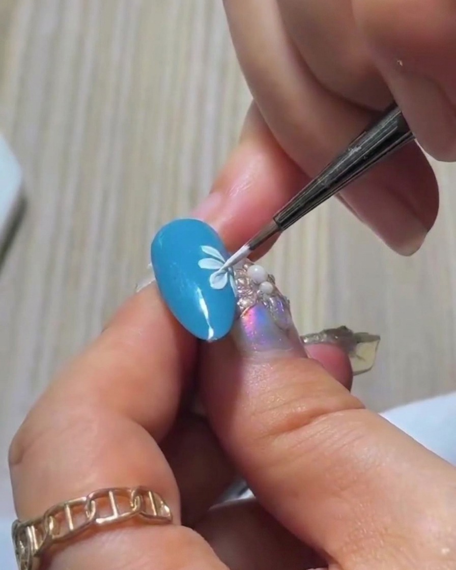 Eine Nahaufnahme des vierten weißen Blütenblatts, das auf den blauen Nagel gemalt wird, wodurch die erste Reihe des Gänseblümchen-Designs aufgebaut wird.