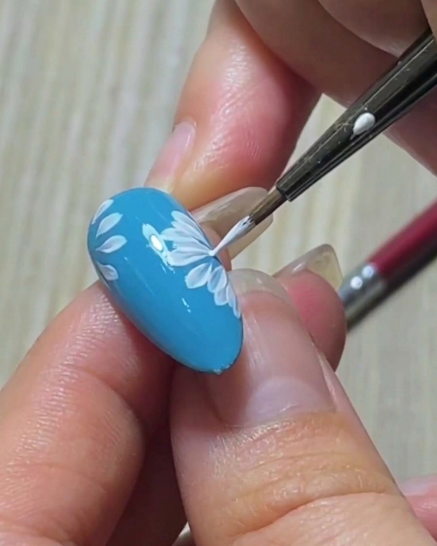 Nahaufnahme eines Liner-Pinsels, der die scharfe Spitze eines weißen Blütenblatts auf einer blauen Nagelbasis verfeinert.