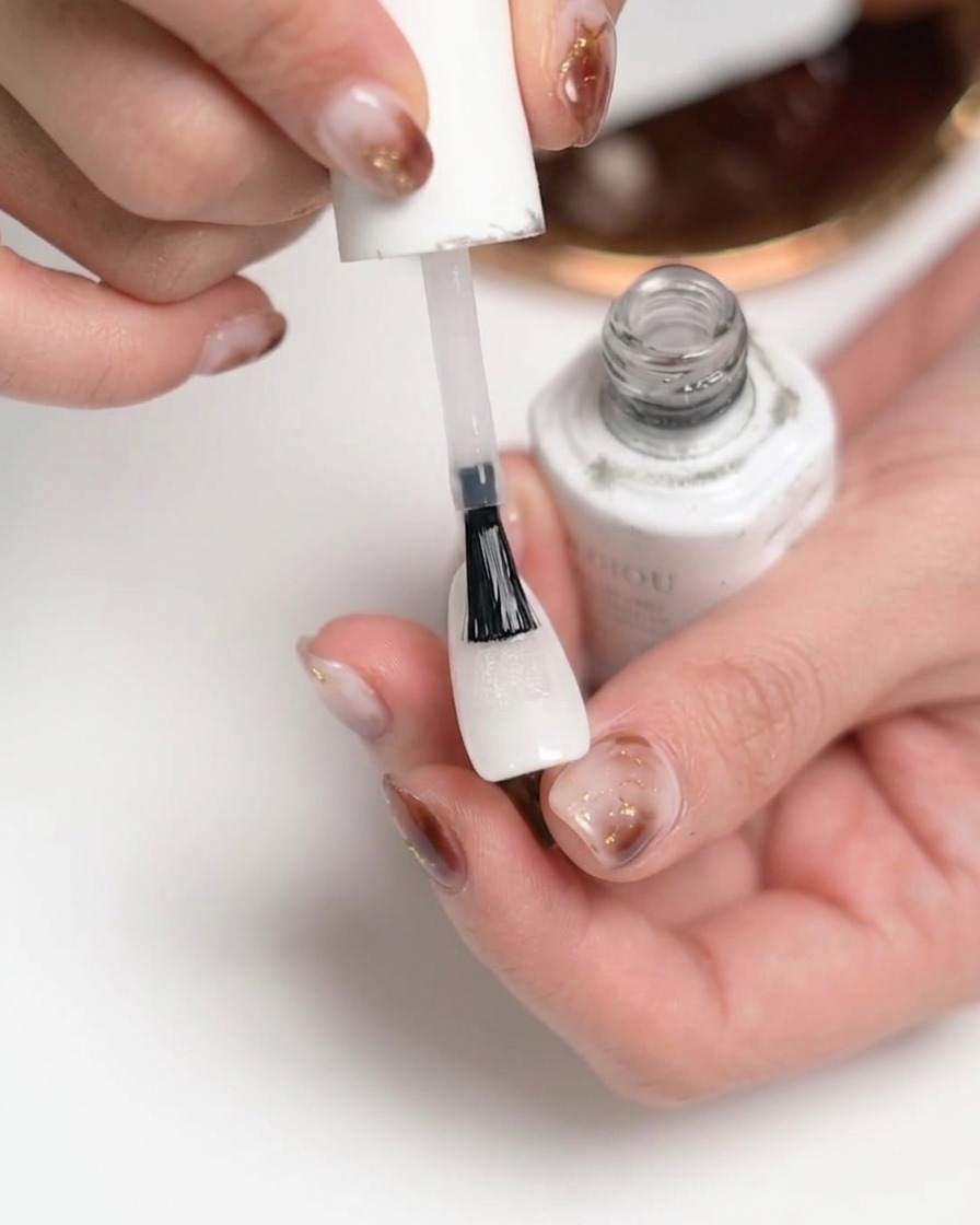 Nahaufnahme einer Hand, die einen klaren, matten Überlack auf einen weiß lackierten Fingernagel aufträgt.