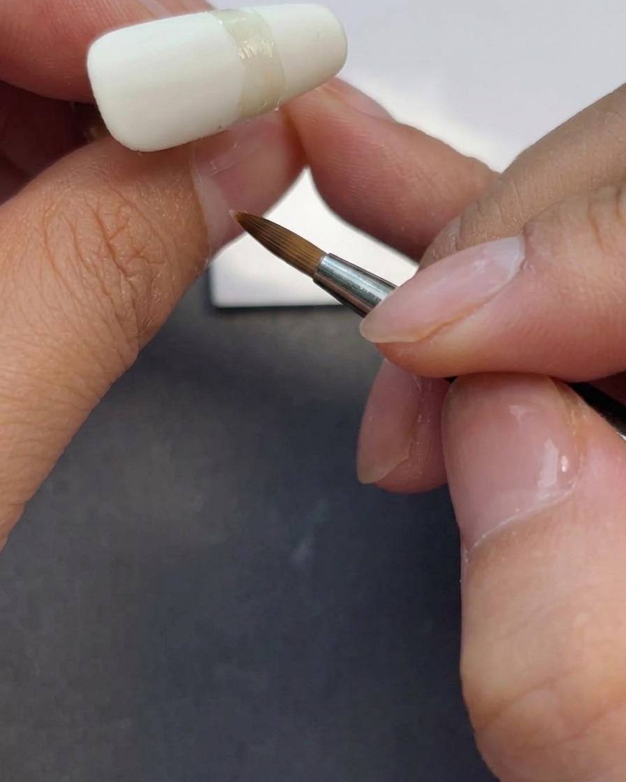 Eine Nageldesignerin trägt mit einem Gel-Pinsel matten Top Coat auf einen milchig-weißen Nagel auf.