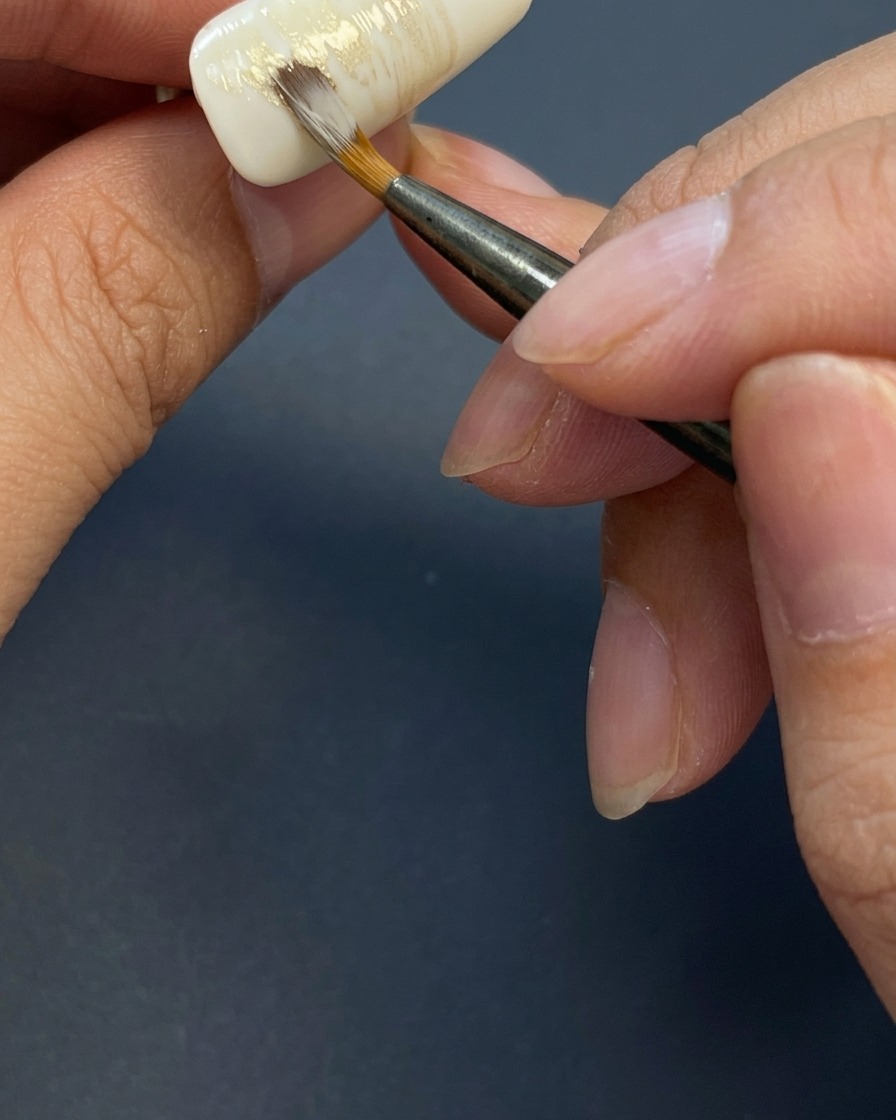 Ein Pinsel zieht leichte, streifige horizontale Striche mit erdigen Gel-Farben auf den weißen Nagel.