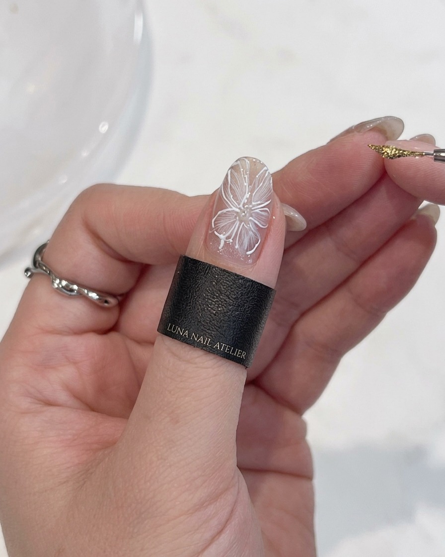 Nahaufnahme einer Hand mit einem feinen Detailpinsel, die einen präzisen Punkt aus metallischem Goldgel in die Mitte einer weißen Blume auf einem Fingernagel setzt.