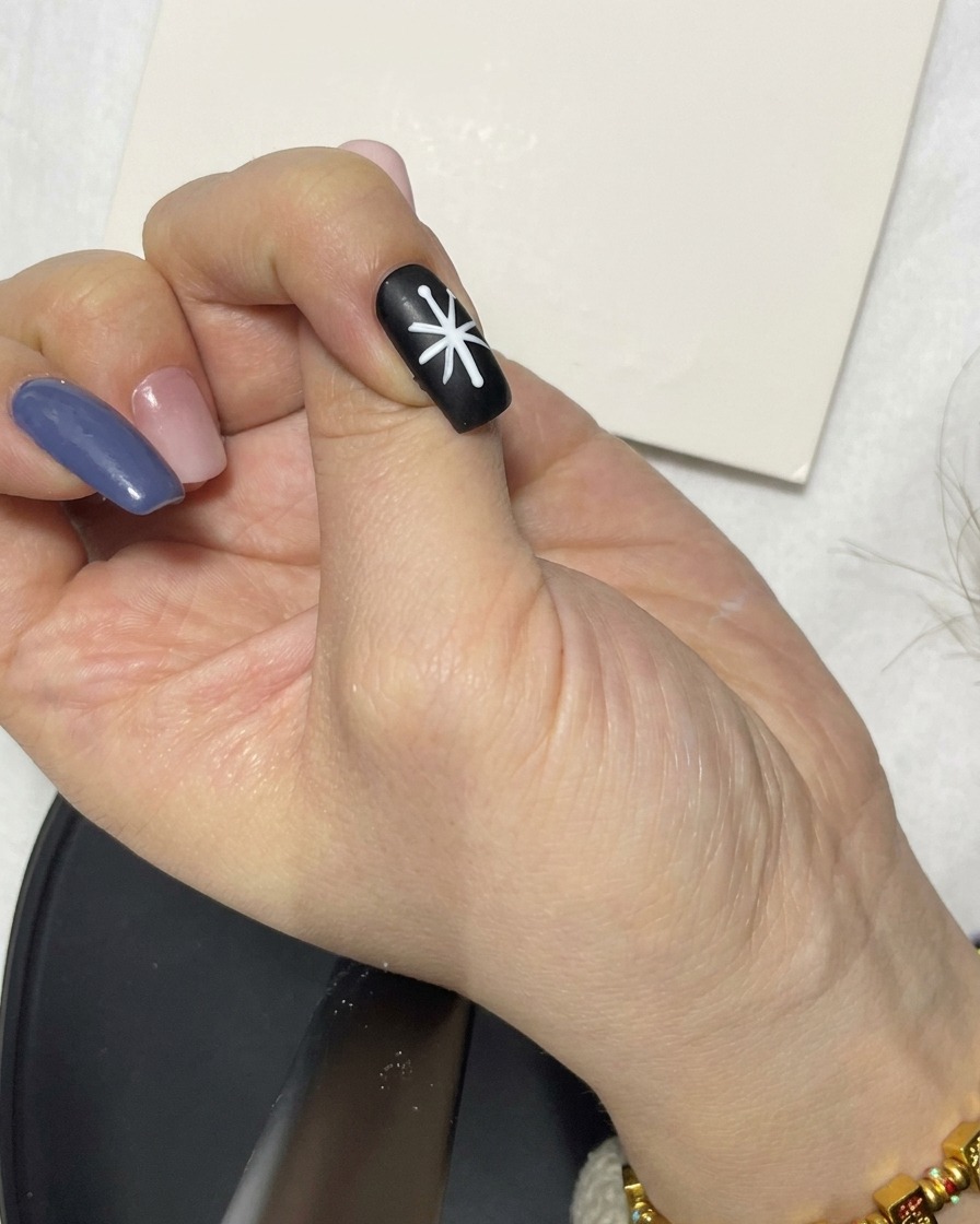Eine Hand, die mit einem Liner-Pinsel die weißen Liniensegmente einer Blumenform auf einem schwarzen Nagel nachzieht.