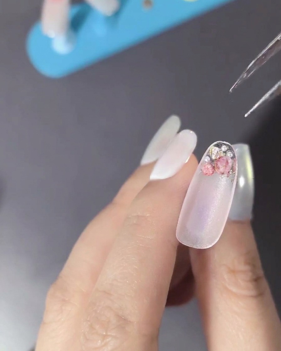 Nahaufnahme eines Nagels, der mit Trockenblumen, Perlen und kleinen metallischen Strasssteinen verziert ist.