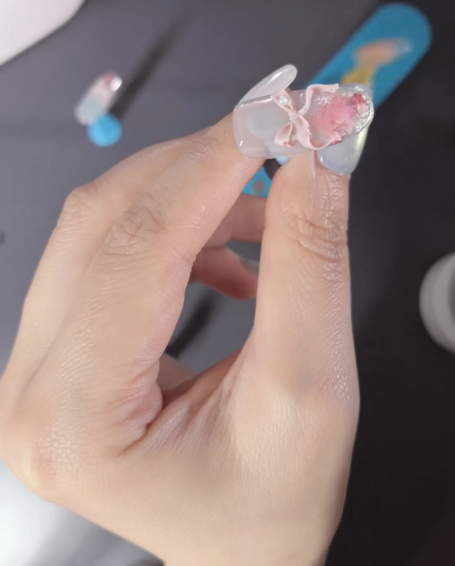 Nahaufnahme eines Fingers mit einer rosafarbenen 3D-Schleife und Perlenakzenten auf dem Nagel.