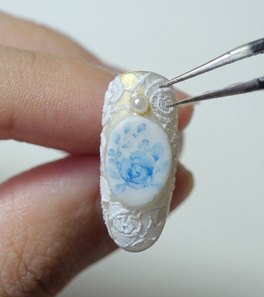Eine Pinzette hält eine kleine weiße Perle über einen Nagel mit blauer Rose und weißer 3D-Spitze.