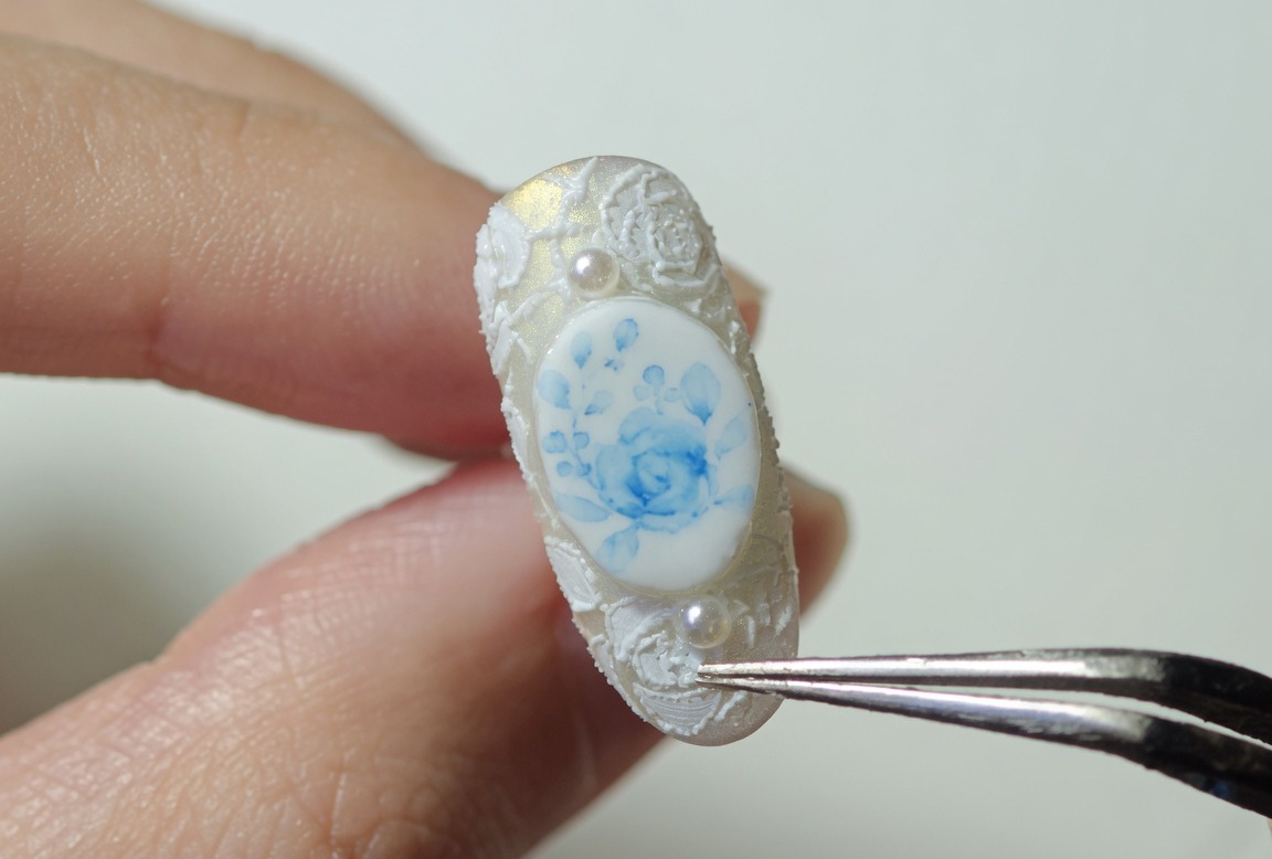 Nahaufnahme eines verzierten Nagels mit blauen Rosen und weißer Spitze, gehalten von einer Pinzette, bereit für die finale Aushärtung.