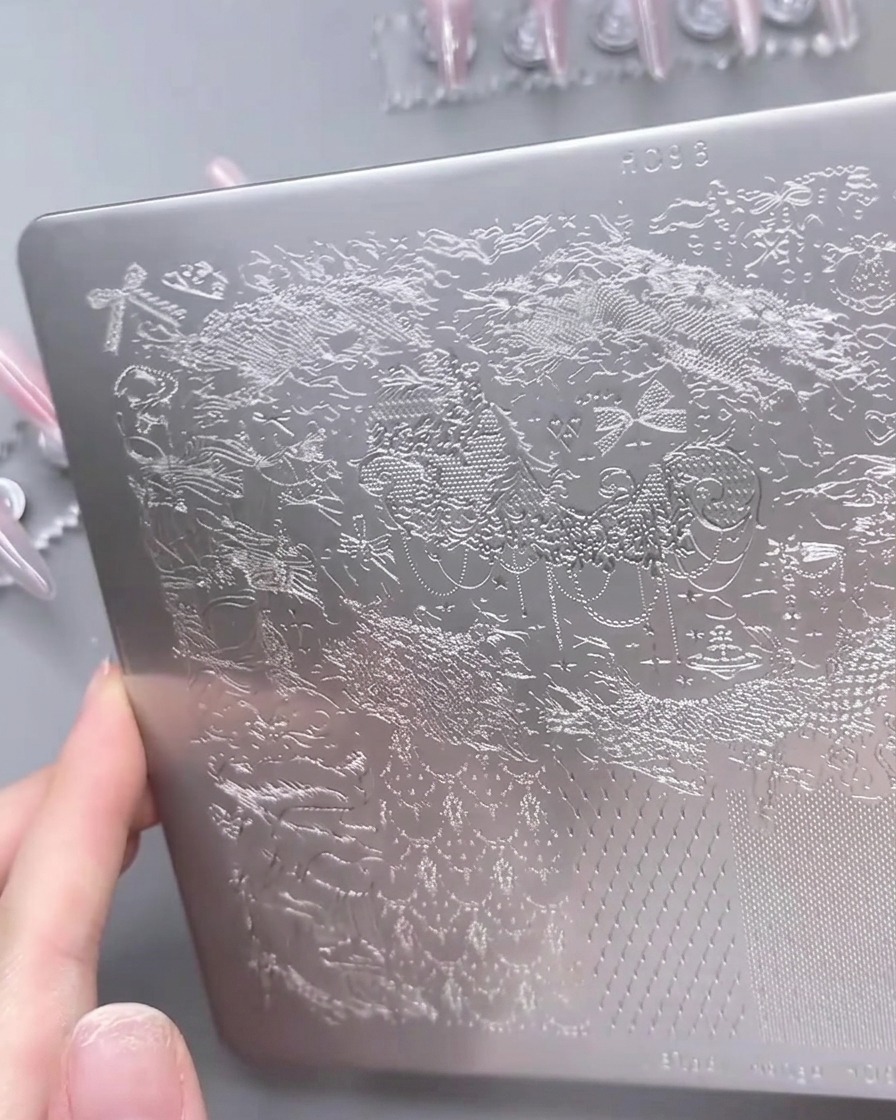Eine Nahaufnahme einer Metall-Stamping-Schablone, auf der weißer Gel-Nagellack mit einem Schaber über ein detailliertes Spitzenmuster verteilt wird.