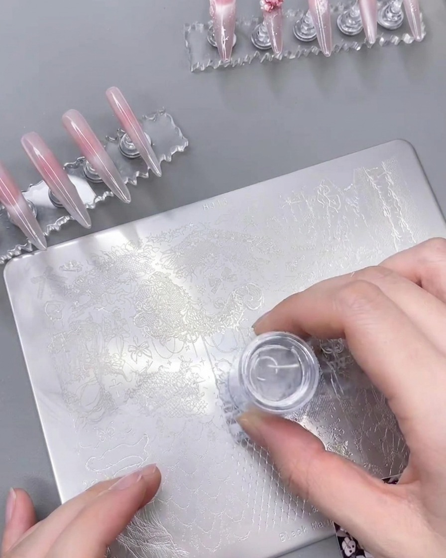 Ein durchsichtiger Silikon-Nagelstempel wird auf eine Stahlschablone gedrückt und nimmt das weiße Spitzen-Design erfolgreich auf.