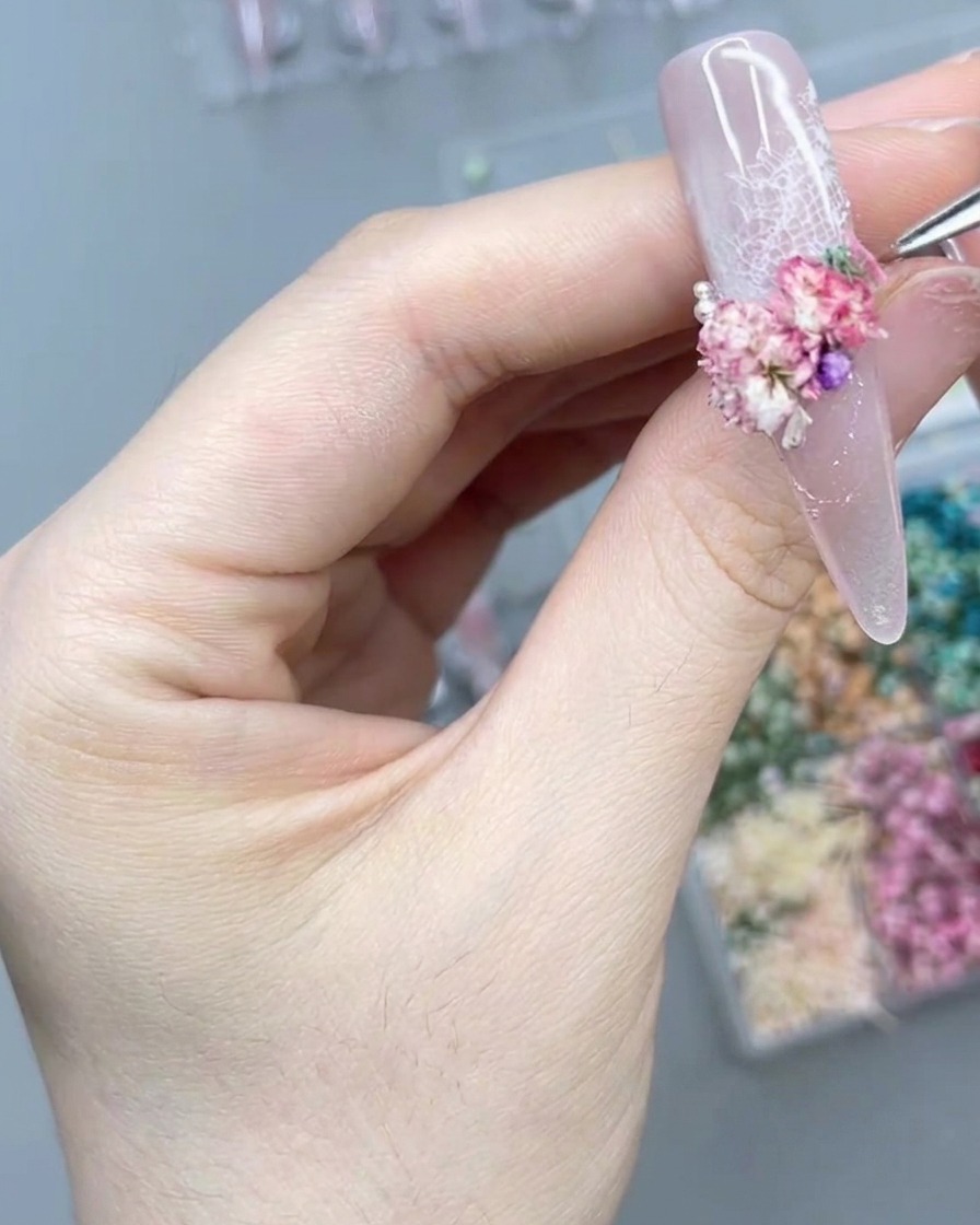 Eine Nahaufnahme einer Hand, die mit einem Präzisionswerkzeug vorsichtig eine winzige Perle neben einer Ansammlung von rosa Trockenblumen auf einem langen Press-On-Nagel platziert.
