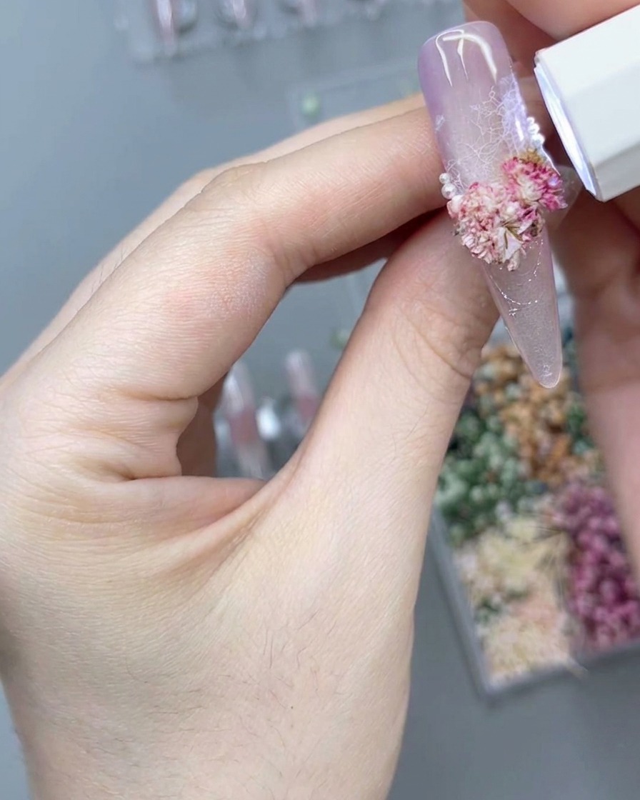 Eine tragbare weiße UV/LED-Lampe strahlt nach unten über ein rosa 3D-Blumenarrangement auf einem langen rosa Press-On-Nagel, um das Design auszuhärten.