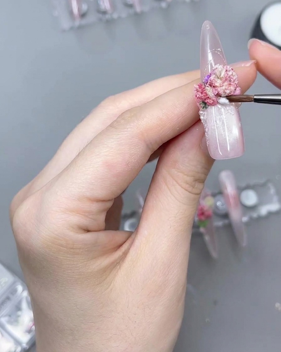 Eine Nahaufnahme eines feinen Detailpinsels, der ein klares Produkt um die Basis einer rosa Blumenansammlung auf einem langen Press-On-Nagel aufträgt.