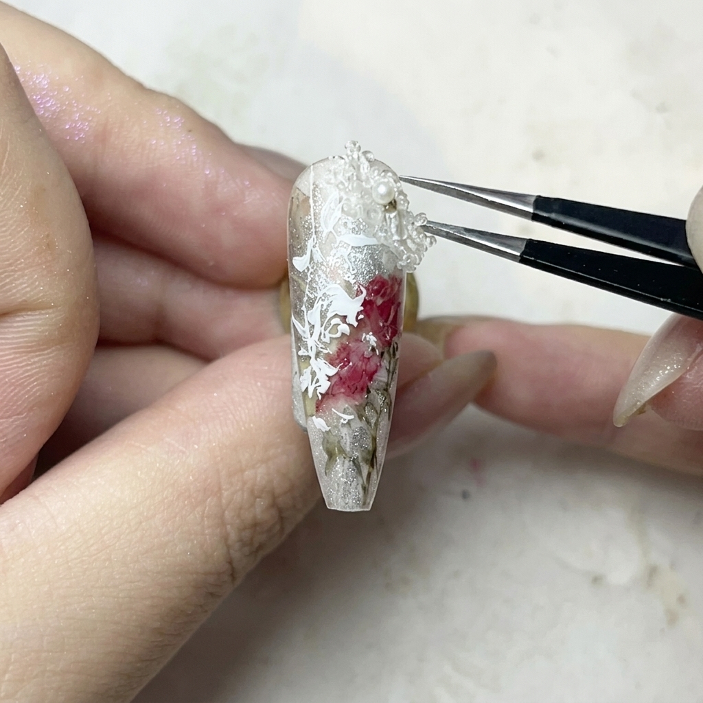 Eine Pinzette platziert vorsichtig einen kleinen, weißen 3D-Blumen-Charm auf einen Punkt klaren Gel-Klebers.