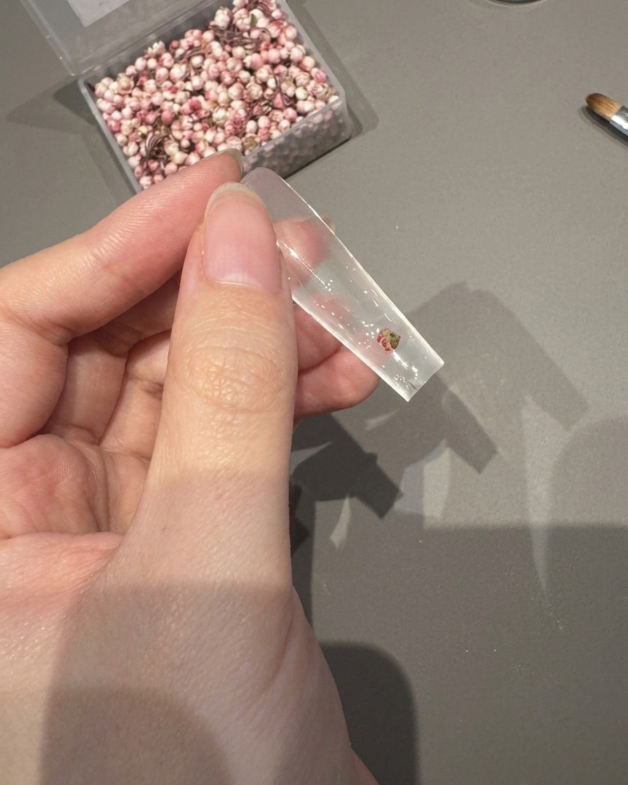 Eine Pinzette platziert eine kleine Trockenblume auf einem mit feuchtem Haftgel bedeckten Nagel-Tip.
