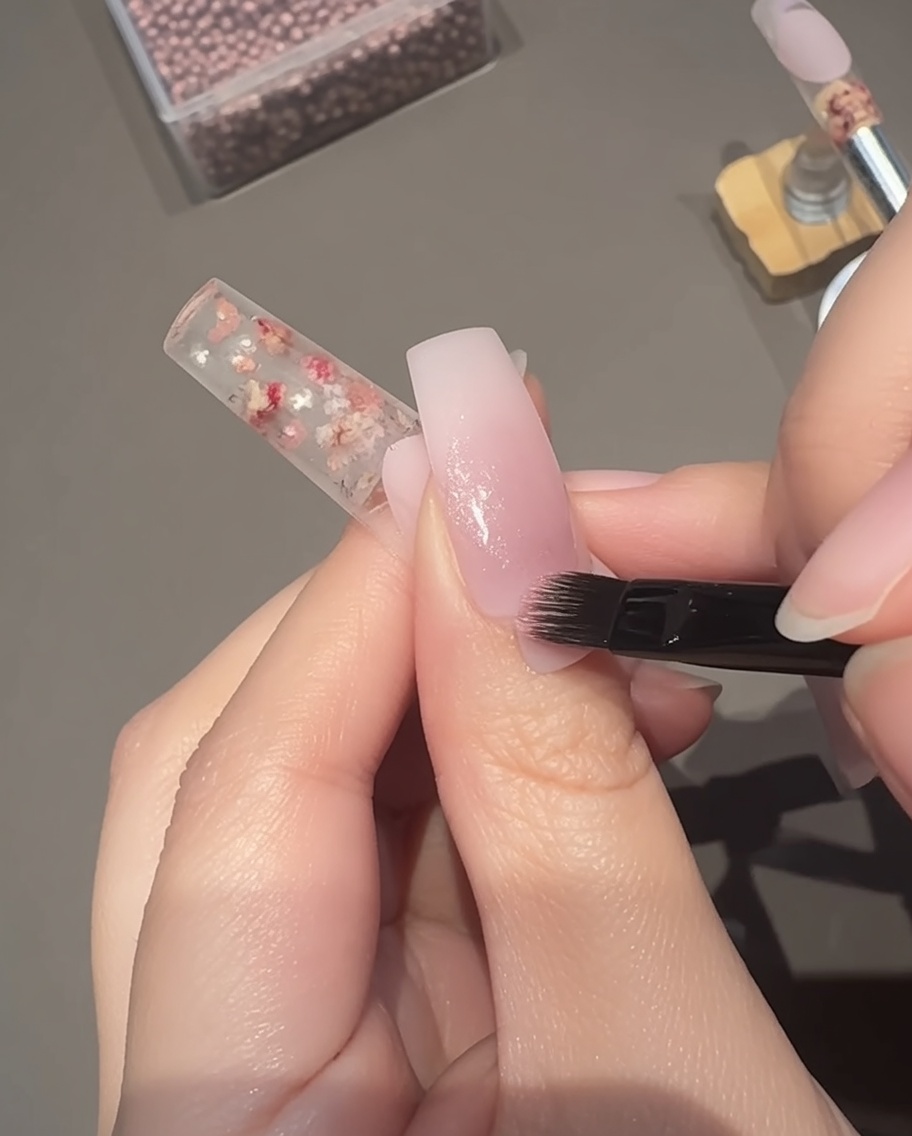 Ein Pinsel trägt ein transparentes rosa magnetisches Cat-Eye-Gel auf das Nagelbett auf.